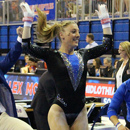 Gymnast in a black and blue leotard with arms raised, smiling after a performance. Blurred spectators in the background.