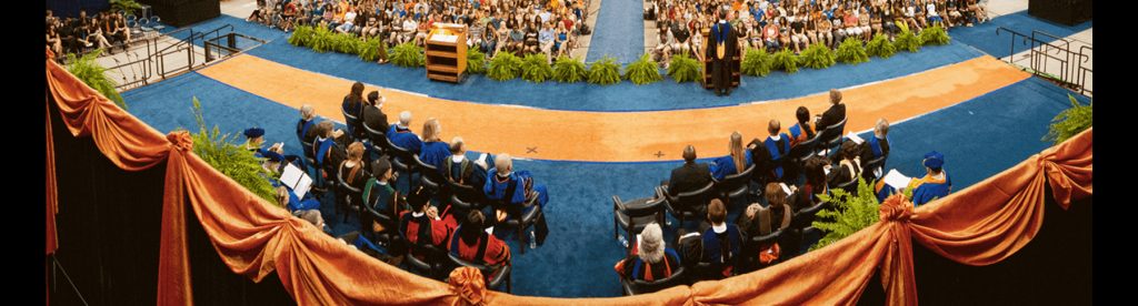 Wide view of a crowded indoor stadium with rows of seated people in academic robes. A stage with a podium is visible in the distance.